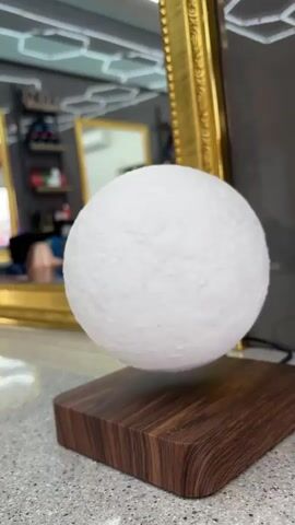 A white, textured sphere resembling the moon hovers above a square wooden base. The base sits on a speckled countertop. Behind the levitating sphere are two mirrors. The larger mirror, framed in gold, reflects a cluttered shelf in the background. The smaller mirror has a geometric light fixture above it. A power cord is visible near the base, presumably powering the levitation device.