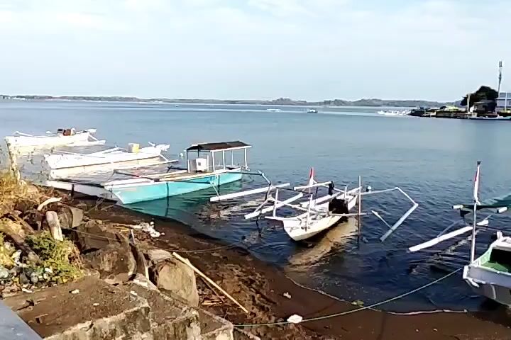 The video shows a shoreline with several boats docked. The water is calm and the sky is clear. The boats are of various sizes and colors, with some appearing to be fishing boats. There are also some structures visible on the shore, including what looks like a small building or shelter. The overall impression is one of a peaceful, coastal scene.
