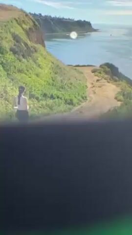 The video depicts a woman jogging on a dirt path along a coastal cliff. The woman has long, dark hair, and she is wearing a white top and dark shorts. The path is surrounded by green vegetation, and the ocean is visible in the background. The sky is blue with some clouds. There is a noticeable glare on the lens.
