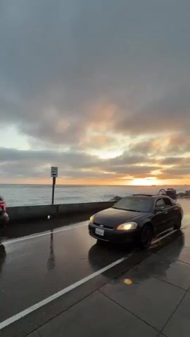 A black sedan with black rims is parked on a wet road next to the ocean. The car has a sunroof open and what appears to be a bicycle rack attached to the trunk with two bicycles on it. A white sign with an arrow pointing left and the words "ONE WAY" is visible on a post to the left of the car. The sky is cloudy with the sun setting, casting a warm orange glow on the horizon and reflecting on the wet road. Several other cars are driving down the road in the distance. The road has a white line marking the lane. The scene appears to be a coastal highway during sunset.