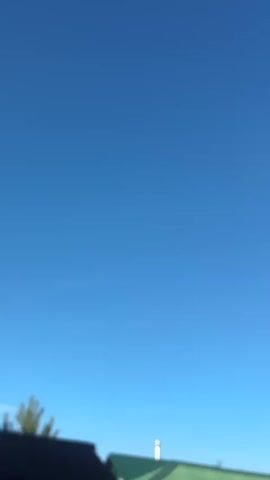 The video shows a bright blue sky with a few blurry green trees and building rooftops visible at the bottom. The camera is tilted upwards and appears to be in motion, possibly handheld, as the image is slightly shaky. No people or specific actions are discernible. The environment suggests an outdoor setting during the daytime.