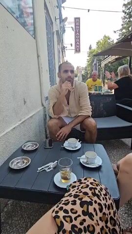 The video shows an outdoor seating area of a restaurant. In the foreground, a person wearing a leopard-print skirt is visible from the waist down, sitting at a black table. On the table are two empty coffee cups on saucers, an ashtray with cigarette butts, a glass with amber liquid, a cell phone, and silverware. A man in a light-colored short-sleeved shirt and white shorts is sitting on a dark-colored couch to the left, looking to the left with his hand on his chin. Behind him, to the right, another man in a yellow shirt is seated at a table with two women. One woman in a black shirt is gesturing with her hands, and the other woman in a yellow top is looking at her phone. Umbrellas provide shade over the seating area. A "RESTAURANT" sign hangs vertically on the building. Trees and other buildings are visible in the background. The lighting suggests it is daytime.