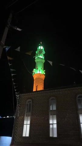This is a nighttime video capturing a minaret illuminated with green and orange lights. The minaret is tall and cylindrical, with intricate brickwork and architectural details, topped by a green, lantern-like structure. It appears to be part of a mosque, with a section of the mosque's roof, featuring arched windows, visible in the foreground. Decorative flags or banners are strung across the scene, adding a festive element. The overall mood is serene and possibly celebratory, given the vibrant lighting. The time of day is clearly night, indicated by the dark sky. While specific weather conditions are not discernible, the clear visibility suggests fair weather. The setting appears to be an urban or semi-urban area, given the presence of electrical lines and streetlights. There is no discernible text in the video.