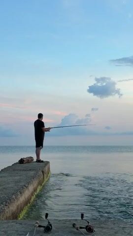 A man stands on a concrete pier and fishes with a long rod. The sea is calm, and the sky is a light blue with pink and white clouds. Two fishing reels are visible in the foreground.