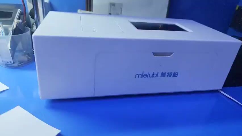 A close-up shot of a white electronic device on a blue surface. The device has a black screen on top and the text "mletubl" and Chinese characters on the front. A hand is visible on the left side of the frame, appearing to interact with the device. In the background, a white office-style desk is cluttered with various items including a pencil holder with pens and a white power strip. The wall behind the desk is painted blue.