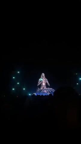 A vertical video captures a concert stage at night. In the center, a large, illuminated sculpture of a person in a seated, meditative pose dominates the frame. The sculpture is covered in reflective material and appears to be projected with cosmic imagery, including galaxies and stars, which shift and change throughout the video. White and blue spotlights emanate from behind the sculpture, illuminating the dark sky and creating dramatic light beams. In the foreground, the silhouettes of a crowd of people are visible, facing the stage. Some bright lights from stage equipment are also visible on the left side of the frame. The overall atmosphere is that of a large-scale musical performance with impressive visual effects.
