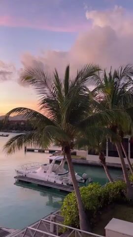 This video shows a serene harbor scene at sunset. A white speedboat is docked in the foreground. Palm trees frame the view on the right side of the image. In the background, several sailboats and other vessels are moored in the calm water. The sky is a vibrant mix of pink, purple, and orange hues, with scattered clouds adding texture. Distant, silhouetted mountains form the horizon. The overall impression is one of tranquility and natural beauty, with a touch of tropical luxury suggested by the docked boats.