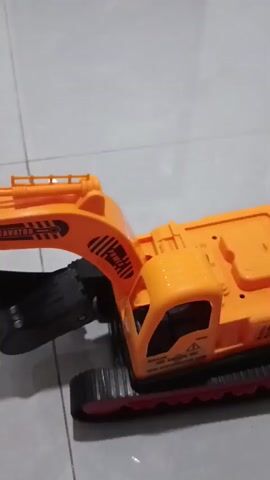 A close-up shot of an orange toy excavator is presented. The excavator is on a tiled surface. The camera is held low, and the excavator is in the foreground. The excavator's arm is extended, and its bucket is tilted downwards. The excavator has black tracks with red accents. The excavator's body is orange, and it has a driver's cabin with black windows. The excavator is stationary for the duration of the video. The lighting is adequate, and the image is slightly blurry due to the motion of the camera.