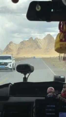 The video captures a first-person view from inside a car, looking out through the windshield at a long, straight asphalt road. In the foreground, the car's dashboard is visible, featuring a device (possibly a radar detector or dashcam) mounted in the center, and two small, decorative figurines, one resembling a laughing Buddha, on the right side. A rearview mirror is present at the top, from which red prayer beads and a yellow pouch adorned with what appears to be Tibetan script are hanging.

A white car approaches the camera vehicle from a distance on the road, eventually passing it. The background is dominated by a range of large, barren, light brown mountains under a cloudy, overcast sky, suggesting a dry or desert-like environment. Faint red text is visible on the side of one of the distant mountains, though it is illegible. On a screen visible on the car's dashboard, text reads "抖音火爆歌曲" (Douyin popular songs) and "2021没意义..." (2021 meaningless...). The scene depicts travel through a mountainous region, likely in the vicinity of Zagor town, China, during the daytime.