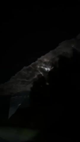The video shows a night sky with the moon visible behind clouds. The clouds are illuminated by the moon and appear to be moving. There are also dark silhouettes of trees in the foreground. The video is short and appears to be a recording of a natural phenomenon.
