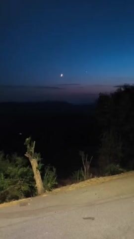 A video shows a dark, rural landscape at dusk or dawn. The sky is a deep blue, transitioning to lighter shades near the horizon, where faint hues of pink and purple are visible. In the upper portion of the sky, two celestial bodies are visible: a crescent moon and a bright star or planet. Below the sky, dark, silhouetted mountains create a jagged line against the horizon. In the foreground, trees and vegetation are dark and indistinct. A paved road or path runs diagonally across the lower part of the frame. On the right side of the frame, the rear of a white camper van is visible, parked on the side of the road. The van has windows and roof-mounted equipment. A second vehicle, possibly another camper van, is partially visible further up the road. The overall impression is one of quiet solitude in a natural setting at the transition between day and night.