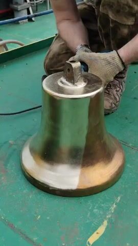 The scene features a person polishing a large, golden-colored bell with a power tool on a green surface, presumably the deck of a ship. The individual is kneeling, wearing camouflage trousers, work gloves, and appears to be focused on cleaning the bell.  The bell is visually split, with one half already polished to a bright sheen and the other retaining a darker, tarnished appearance. A power cord snakes across the green surface, indicating the tool is electrically powered. The background shows parts of the ship's structure and deck equipment.