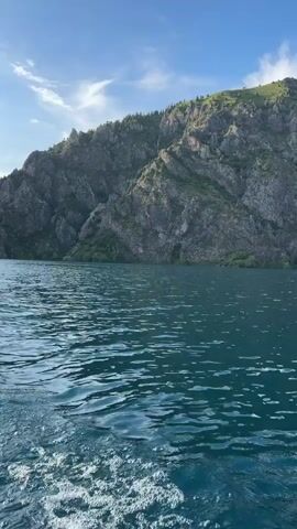 A video taken from a boat on a body of water shows a mountain with trees on its slopes. The water is choppy, and the sky is blue with some clouds. The camera is moving, as evidenced by the wake behind the boat.