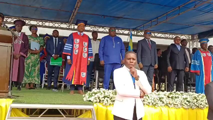 On an outdoor stage, numerous people stand together. At the forefront, a person in a white blazer holds a microphone and appears to be speaking. Behind them, several individuals are dressed in formal attire, including suits and academic robes. A Congolese flag is visible in the background. The stage is decorated with white floral arrangements. The scene appears to be a formal event, possibly a graduation ceremony or an official gathering.
