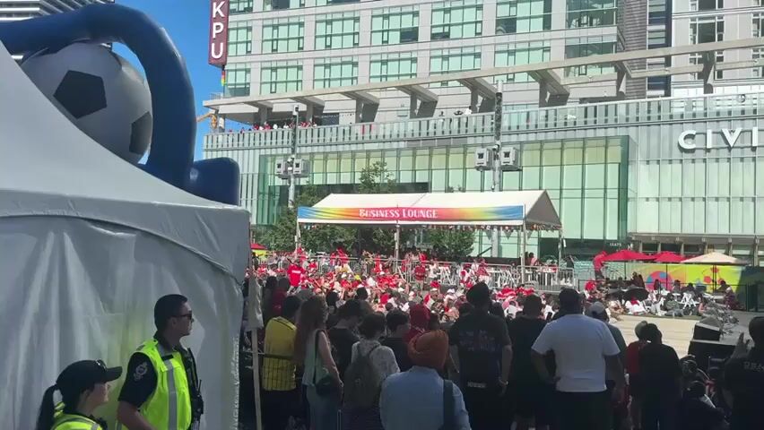A large crowd of people are gathered outdoors, many wearing red clothing. There is a white tent in the foreground to the left with two security guards standing in front of it. Behind the tent, a large inflatable soccer ball is visible. In the background, a stage is set up with a banner that reads "BUSINESS LOUNGE" in rainbow colors. Flags from various countries are displayed along the top of the stage. People are seated and standing around the stage, some appearing to be watching an event or performance. A modern building with a glass facade is visible behind the stage. The overall scene appears to be a public event or festival on a sunny day.