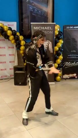The video shows a person dressed as Michael Jackson performing a dance routine in front of a banner that says "MICHAEL JACKSON". The person is wearing a black sequined jacket, black pants with white stripes, and white socks. They are also wearing black shoes and gloves. The background includes a movie poster, balloons, and a speaker. The person is performing the iconic "moonwalk" dance. The video is shot from a low angle and the lighting is dim.