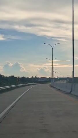 A drone shot pans across a highway at sunset. The sky is filled with soft, white clouds against a blue background, with the sun setting on the right side of the frame, casting a warm glow. The highway itself is made of concrete, with a white line marking the edge on the left and a yellow line on the right. A concrete barrier runs along the right side of the road, and several streetlights with curved poles are spaced evenly apart. On the left side of the highway, there are trees and a few buildings in the distance, with a curved road sign indicating a turn. The overall scene is serene and captures the beauty of a sunset on an open road.