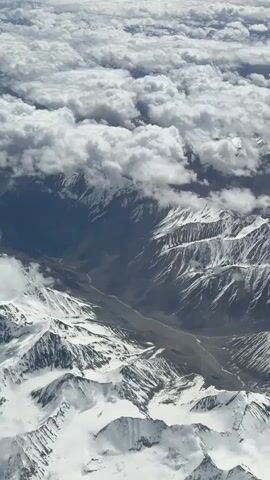 An aerial view of snow-capped mountains below a thick layer of white clouds. The mountains are rugged and appear to be a valley between them. The scene is lit by natural sunlight, creating shadows on the mountains. The camera is slowly panning downwards and slightly to the left.