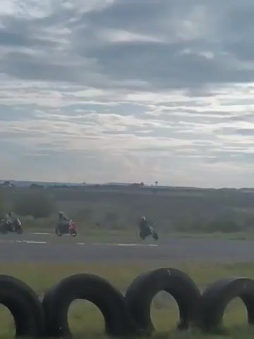 The video shows four motorcycles racing around an outdoor track on a cloudy day. The track is lined with stacked tires. The horizon includes a flat, grassy landscape with trees in the distance, and the sky is partly covered with white clouds.