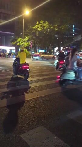 This is a nighttime, first-person perspective video of a busy street intersection. The camera is moving and appears to be mounted on a vehicle. There are several electric scooters and bicycles on the road, with people riding them. In the foreground, there are two white electric scooters. In the middle ground, there are several people on various electric scooters and bicycles, crossing the street or waiting at the intersection. There are also some cars in the background. The street is wet, indicating it may have recently rained. Trees and buildings line the street, and there are traffic lights and streetlights visible. The overall impression is of a lively urban environment at night.