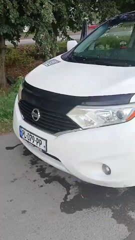 The video shows a white minivan parked outdoors on a paved surface. The minivan is a Nissan, indicated by the logo on the grille. The license plate is visible and reads "PL-079-PP".

The side of the minivan features prominent text and a QR code. The text reads "TOUR IN GEORGIA" and "Transfer", followed by two phone numbers: "+995 579 77 05 57" and "+995 593 46 86 80". A WhatsApp logo is also present next to the first phone number. A square QR code is affixed to the rear panel, labeled "MINIVAN SERVICE".

The weather appears to be overcast, with clouds reflected in the windows of the vehicle. The surrounding environment includes greenery, with trees visible in the background. The time of day is likely daytime. The setting is consistent with a roadside or parking area, possibly in a town or city, given the paved surface and nearby trees. The overall impression is of a vehicle used for transportation services, specifically tours and transfers in Georgia.