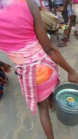 This video captures a bustling market scene, likely in Yenegoa, Nigeria, given the context. The overall impression is one of lively commerce and community interaction.

The subjects are predominantly people, with a mix of men and women, many of whom appear to be vendors or shoppers. They are dressed in a variety of clothing, including traditional African attire, patterned skirts, and casual wear. Some individuals are seen wearing hats, possibly for sun protection.

Objects in the scene include market stalls, umbrellas of various colors (pink, yellow, blue) providing shade, buckets, baskets, and what appear to be agricultural products like brooms or bundles of straw. A wheelbarrow is also visible, suggesting the transport of goods. The ground is a rough, earthy surface, typical of an outdoor market. In the background, there are glimpses of vehicles, including a car and a white van, indicating the market's accessibility by road.

The activity taking place is the operation of an outdoor market. People are moving around, browsing, and interacting. The camera focuses on several individuals, some of whom seem to be engaged in selling or preparing to sell items. The overall atmosphere appears energetic and busy.

Notable details include the bright, overcast sky, suggesting it's daytime, possibly late morning or afternoon. The presence of umbrellas indicates a sunny or potentially rainy climate. The camera work is dynamic, with quick pans and zooms, adding to the sense of lively activity. While specific emotions are hard to discern due to the video quality, the general atmosphere seems positive and engaged. There is no visible text in the provided frames.