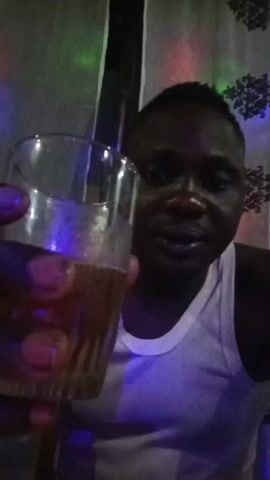 A man in a white tank top is holding up a glass filled with a brown liquid. He appears to be inside, possibly at a party, as there are colored lights flashing in the background. He is looking directly at the camera and talking, possibly making a toast.