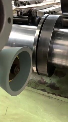 A close-up shot shows a large metal cylinder with a silver, polished surface. A green, cone-shaped roller with a yellow center is positioned next to the cylinder. The roller appears to be in contact with or very close to the cylinder's surface. In the background, a piece of machinery with visible screws and metal components is out of focus. The ground beneath the machinery is a green, painted surface with some dark stains. The lighting is bright, casting reflections on the metal surfaces.