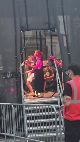 This media file captures a live music performance, likely outdoors given the stage setup and the presence of metal crowd barriers in the foreground. The scene is viewed from behind the barriers, looking up at a stage.

The central figures on stage are musicians. In the foreground of the stage, a woman wearing a dark-colored dress and a pink vest is holding a guitar and appears to be singing or interacting with the audience. To her right, another musician, a man with a shaved head wearing sunglasses and a dark t-shirt and shorts, is playing a guitar. Behind them, a person is seated at what appears to be a keyboard or synthesizer. More musicians and equipment are visible further back on the stage, including at least one other guitar player. The stage is lit with vibrant pink and red lights, creating a dynamic and energetic atmosphere.

The setting appears to be urban, with buildings visible in the background through the stage rigging. The time of day seems to be evening or late afternoon, as indicated by the lighting and the need for stage lighting. The overall mood conveyed is one of lively musical entertainment. There is no visible text in the media file.