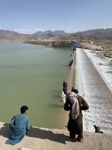 The video captures a wide, sunny, outdoor scene featuring a dam or weir with people crossing it on foot. In the foreground, two men are visible on a dry, sandy bank. One man, wearing a blue tunic and trousers, is seated on the left, facing away from the camera and looking towards the water structure. To his right, another man stands, also facing away from the camera, observing the dam. He wears dark traditional clothing, with a light-colored shawl draped over his left shoulder and a striped cap on his head.

The main focus is a large concrete or stone dam/weir that stretches horizontally across the frame. On the left side of the structure, there is a calm, wide body of greenish-brown water, resembling a reservoir or a large river. On the right side of the structure, water is actively flowing over its edge, creating a cascade of white, foamy, turbulent water that rushes downwards.

A line of approximately 7 to 8 individuals is seen carefully walking across the top surface of the dam from right to left. They are dressed in traditional attire and appear to be wading through shallow water that covers the dam's surface, while simultaneously navigating the flow as water spills over the edge.

In the mid-distance across the reservoir, a blue rectangular sign is visible on the far bank, but its text is illegible due to the distance. The background consists of a vast expanse of dry, rugged, and barren mountains under a clear, bright blue sky, suggesting a sunny day in an arid or semi-arid region. The overall scene depicts a functional crossing point over a significant body of water in a seemingly remote and natural landscape.