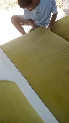 A person kneels on the ground and uses a sponge to scrub a large, olive-green couch cushion. The couch appears to be outdoors, with dirt and gravel visible in the background. The person is wearing a light blue t-shirt and khaki shorts.
