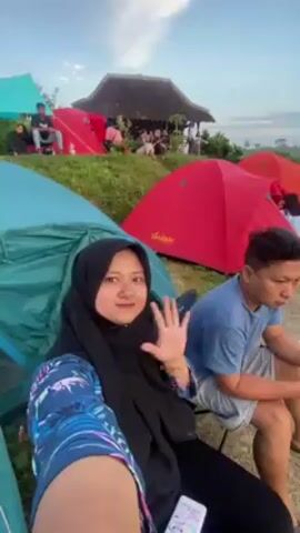 The video shows a camping site on a hill overlooking a bay. Several tents are set up, including a blue, a red, and an orange one. People are seen sitting and standing around the tents. One woman wearing a black hijab waves to the camera. The camera pans to show the view of a bay with a visible sunset in the sky. The landscape is lush and green. A man in a blue shirt is sitting near the tents, looking at his phone. The overall atmosphere is relaxed and scenic.