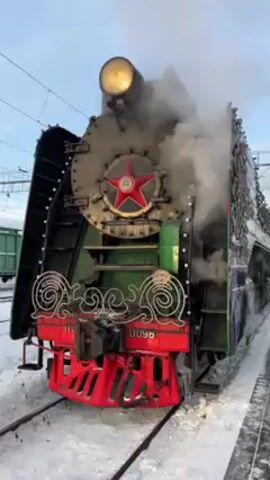 A steam locomotive stands in a snow-covered train yard. The locomotive is painted in a variety of colors, including black, silver, green, and red. It is decorated with a red star on the front and a light on top. Steam is coming out of the top of the locomotive, and there is a green train car in the background.