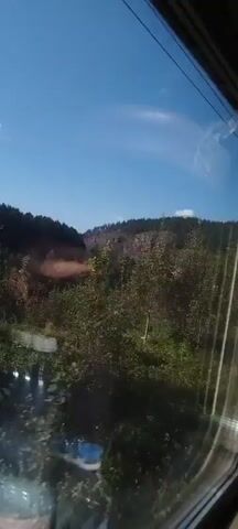 The video shows a view from a moving train or vehicle, looking out of a window. The scene outside is a rural landscape with rolling hills covered in trees and vegetation. The sky is clear and blue. There are some signs of human activity, such as what appears to be a power line pole and possibly a road or path winding through the landscape. The light suggests it is daytime, possibly mid-morning or afternoon. The video is shot from a slightly low angle, looking upwards at the landscape. There are no people visible in the video. The overall impression is of a scenic journey through the countryside.