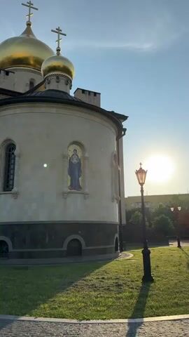 The video shows a side view of an Orthodox church with golden domes against a bright blue sky. The sun is setting or rising, casting a bright glare and long shadows. The church is made of light-colored stone with a darker stone base. There are two large golden domes visible, topped with crosses, and a smaller dome on a side structure. A mosaic icon of a religious figure in blue and red is affixed to the curved wall of the church. There are arched windows and openings at the base of the church. In the foreground, there is a cobblestone path and a well-maintained lawn. A black lamppost with an ornate design stands on the right side of the frame, casting a shadow. A small brown object, possibly a brick or stone, is on the grass in the lower left corner. The environment appears to be a quiet, outdoor space. The camera pans slightly, changing the perspective of the church and its surroundings. There are no people visible in the video.