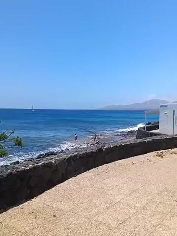 The video presents a bright, sunny coastal scene in Puerto Del Carmen, Spain. In the foreground, a light-colored, sandy-textured paved path occupies the lower half of the frame, separated from the sea by a low, dark stone wall constructed from irregular blocks.

Beyond the wall, a rocky shoreline meets the clear, blue-green waters of the ocean. Gentle waves can be seen breaking against the shore. Two individuals are visible wading in the shallow water near the rocks, seemingly enjoying the sea.

In the mid-distance, a lone sailboat is discernible on the horizon, sailing across the calm, deep blue ocean. Further in the background, a line of distant mountains stretches across the landscape under a vast, clear, cloudless blue sky. To the far right, a small white building or shelter with a light-colored canopy is partially visible, blending into the bright surroundings. A patch of green foliage from a bush or tree can be seen on the left side of the frame, adding a touch of greenery to the otherwise stark coastline. The overall atmosphere is peaceful and serene, indicative of a pleasant day by the sea. No discernible text is visible.