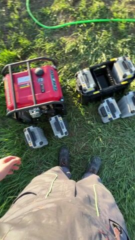 Here's a concise description of the video:

The video shows a field with a worker preparing to use a large drone, likely for agricultural purposes.  A red generator provides power. The worker is visible briefly next to the drone, appearing to check or adjust it. The drone has multiple propellers and a liquid tank attached. Several gray battery charging units are on the ground near the generator and a container for batteries. A white pickup truck is pulling a small trailer is nearby on a dirt road. The environment is rural, with a field of low-lying plants, grass, and scattered trees. The sky is blue with some clouds.