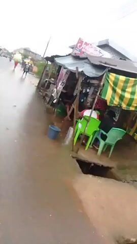 A first-person perspective video shows a wet, muddy street with small roadside shops. A person is walking and filming, with the camera moving. To the right, there are several makeshift stalls with corrugated metal roofs supported by wooden poles. The stalls are filled with various items, some of which are covered by tarps. Several people are seated at the stalls. On the left side of the street, there are more shops and people, some on motorcycles and others walking. A large poster of a woman with long black hair is displayed on one of the stalls. There is a hole in the ground near the front of one of the stalls. The sky is overcast and the street appears to have recently rained or is still experiencing light rain.