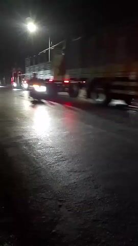 A nighttime video shows a street scene with vehicles and buildings. A large truck with its brake lights illuminated is visible on the right side of the frame. On the left, several motorcycles are passing by, their headlights cutting through the darkness. The road appears wet, reflecting the lights. In the background, there are storefronts with lights and signage. One building has a prominent sign that reads "TIKI". Several parked motorcycles are also visible in front of one of the establishments. A tuk-tuk also passes through the scene. The overall atmosphere is dark with intermittent artificial lighting.