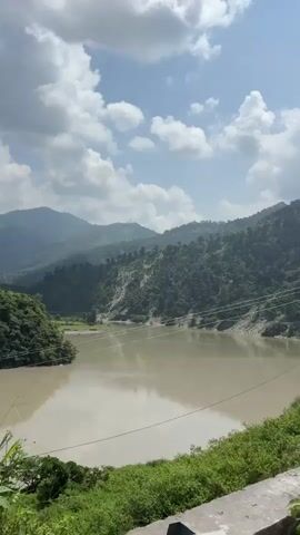 The video begins with a wide shot of a murky body of water, possibly a lake or a slow-moving river, flanked by lush green hills. The sky is partly cloudy, with fluffy white clouds scattered against a blue backdrop. As the video progresses, the camera pans slightly to the right, revealing more of the mountainous terrain. Electrical poles with wires are visible in the mid-ground, strung across the landscape. The foreground shows a concrete barrier and some vegetation, suggesting the footage was taken from a roadside perspective. The overall scene depicts a serene, natural environment, possibly in a hilly or mountainous region.