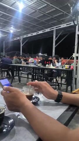 The video shows a group of young men gathered at an outdoor cafe in Kutapadang, Indonesia, at night. The setting appears to be casual and lively, with multiple tables and chairs under a metal roof structure illuminated by bright overhead lights. A television screen is visible in the background, displaying what looks like a sports broadcast.

In the foreground, a person's hands are visible holding a smartphone and a glass of dark liquid, likely coffee, on a saucer. The person is also holding another glass of water. In the middle ground, several young men are seated at tables, some interacting with each other. Two men in particular are the focus of the camera's attention as they embrace playfully, one with his arm around the other. One of these men is wearing a black t-shirt with the word "Transworld" printed on the back in white lettering.

The overall mood of the scene seems to be relaxed and social, suggesting friends enjoying a night out. The time of day is clearly evening or night due to the artificial lighting and the dark sky. There are no discernible weather cues. The presence of the cafe, the specific signage, and the general ambiance suggest a location in Indonesia, consistent with the provided context.