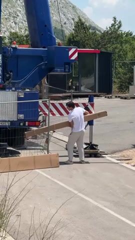Two men are working near a blue crane. One man, wearing a white t-shirt and light-colored pants, is holding a long cardboard box. He is positioned between the crane and a chain-link fence, and appears to be moving the box. Another man is partially visible behind him, also near the crane. The crane has a large blue boom and a red and white striped warning sign. The background shows a wooded area and a mountain. The ground is paved, suggesting an outdoor work site.