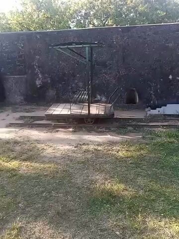 The video shows a grassy area adjacent to an old stone wall. Positioned in front of the wall is a small railcar on a set of tracks.  A woman with dark hair and a cap walks from left to right past the railcar.  She is wearing patterned pants and a black top. Following behind her is a man with a baseball cap, a red backpack, shorts, and a collared shirt. He walks past the railcar to the right. The background shows trees visible above the wall.  The lighting suggests it is daytime.