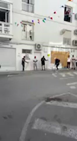 This video captures a street scene during a daytime event, likely a "running of the bulls" or a similar festivity, in what appears to be Vejer de la Frontera, Spain, given the architectural style and provided context.

The scene is set on a narrow street lined with multi-story, white buildings featuring balconies and windows. Colorful bunting hangs across the street between buildings, indicating a festive atmosphere. The sky is bright blue and clear, suggesting sunny weather.

Numerous people are gathered, both on the street and looking out from windows and balconies of the buildings. On the street, individuals are seen moving, some appearing to be participants or observers of the event, while others are swiftly getting out of the way. An animal, resembling a bull or calf, is prominently featured, running down the street. People on the ground react to its movement, with some running or positioning themselves along the side of the street. Spectators are visible in windows and on balconies, watching the action unfold below.

The overall impression is one of a lively public event, characterized by movement and observation, centered around the running animal. No specific text is clearly visible in the video.