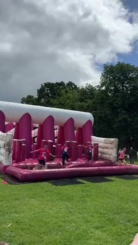 A group of people are on a large inflatable obstacle course outdoors. The obstacle course is pink and white and has a series of poles and platforms. Several people are actively navigating the course, jumping and running. The background shows green grass, trees, and a cloudy sky.