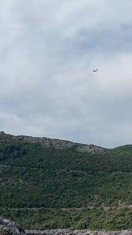 The video captures a wide shot of a scenic landscape under a cloudy sky. In the foreground, the rocky coastline meets the calm, blue water of the sea. Lush green hills rise steeply behind the coastline, covered in dense foliage. A small, dark-colored airplane is visible in the upper portion of the frame, soaring high in the sky. The overall mood is tranquil and picturesque, suggesting a peaceful day outdoors, possibly in the Dubraljevina area of Montenegro, given the location context. The lighting suggests it is daytime, with diffused light due to the cloud cover. There is no visible text in the media.