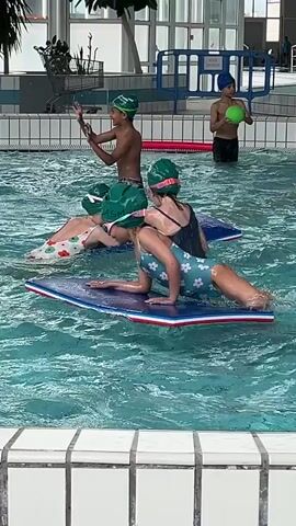 This video shows a group of children engaged in swimming activities within an indoor swimming pool. The setting is bright and well-lit, suggesting daytime. The pool has white tiled edges and clear blue water, indicating a well-maintained facility. The location is Andrézieux-Bouthéon, France.

There are approximately five to six children visible, all wearing swimsuits and swim caps, with some also wearing goggles. Most of the children appear to be between 5 and 10 years old.

In the foreground, a girl wearing a light blue and white patterned swimsuit and a green swim cap with the text "SAINT-ÉTIENNE MÉTROPOLE" is lying on a blue kickboard, propelling herself through the water with her legs. Another girl, also in a green swim cap and dark swimsuit, is standing near her, splashing.

In the background, other children are visible. One boy in a blue swim cap and goggles is holding a pink ball, while another boy in a blue swim cap is holding a green ball. They appear to be playing or practicing with the balls. The children seem to be enjoying themselves, with smiles visible on some faces, indicating a fun and engaging swimming lesson or recreational activity. The overall atmosphere is lively and cheerful.