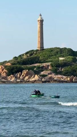 A drone shot shows a lighthouse on a grassy hill overlooking the ocean. Two people in a small boat row through the water in front of the hill. The boat is green and has a small white object on the stern. Another, smaller green boat is tethered to the first boat. The water is blue and choppy. The sky is clear and blue. The sun is shining.