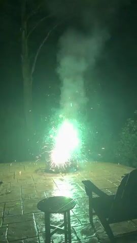 The video shows a short display of fireworks in a backyard at night. The fireworks are set off from a fire pit located on a stone patio. Two small wooden stools and an Adirondack chair are visible on the patio. The fireworks produce sparks and bursts of light in various colors, including orange, white, and green, accompanied by smoke. The patio appears wet, possibly from rain. The background is dark, indicating nighttime. The display lasts for a few seconds, with multiple fireworks going off in succession.