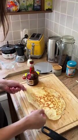 A person's hands are shown over a crepe on a wooden cutting board. The crepe is golden brown with darker brown marks from cooking. A bottle of Grand Marnier liqueur is on the cutting board next to the crepe. In the background, a yellow toaster and various kitchen appliances are visible on a countertop. The person appears to be preparing the crepe, possibly for a dessert.